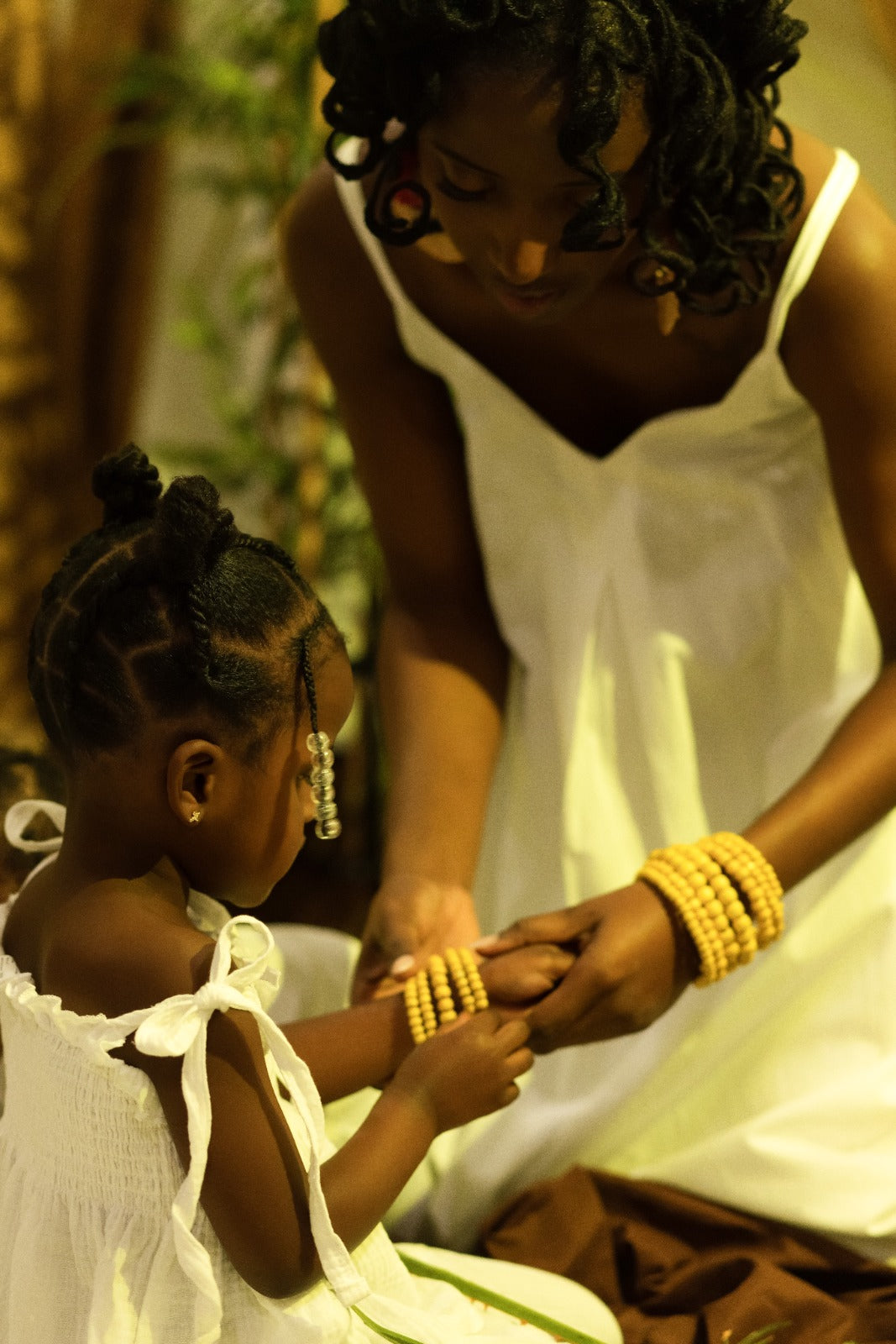 Woman in a white dress sitting with a young girl in a white dress, both wearing yellow bracelets.